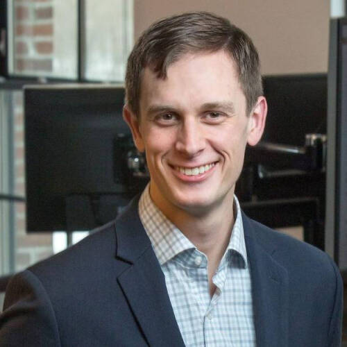 A man stares straight to the camera wearing a gray blazer and a  checked shirt